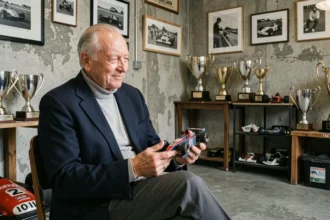 Ancien mentor de Formule 1 assis dans un garage historique tenant un modèle réduit de voiture de course entouré de trophées et de photographies en noir et blanc